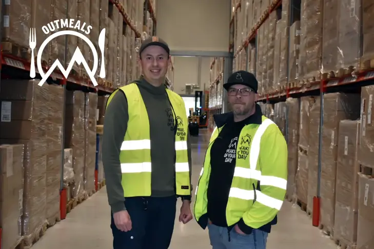 Två män i reflexvästar står i en lagergång med Outmeals-logotypen uppe till vänster.